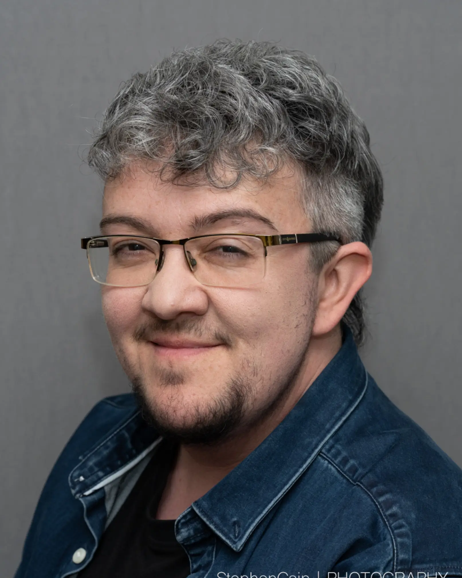 K.C. is a 36-year-old man with silver hair in a curly mohawk/mullet style, with glasses, brown eyes, and a small black beard and moustache. He is smiling for his headshot and wears a black t-shirt and a denim overshirt.