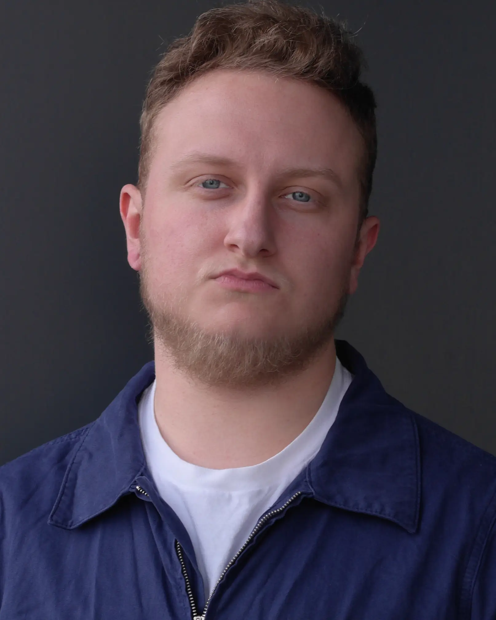 A headshot of the actor Lil J. Bush. They're white with blue eyes, a vaguely warm neutral expression, a jar-shaped head, short dark blonde hair, and a delightful beard. They're wearing a dark blue zip-up collared shirt over a white t-shirt.