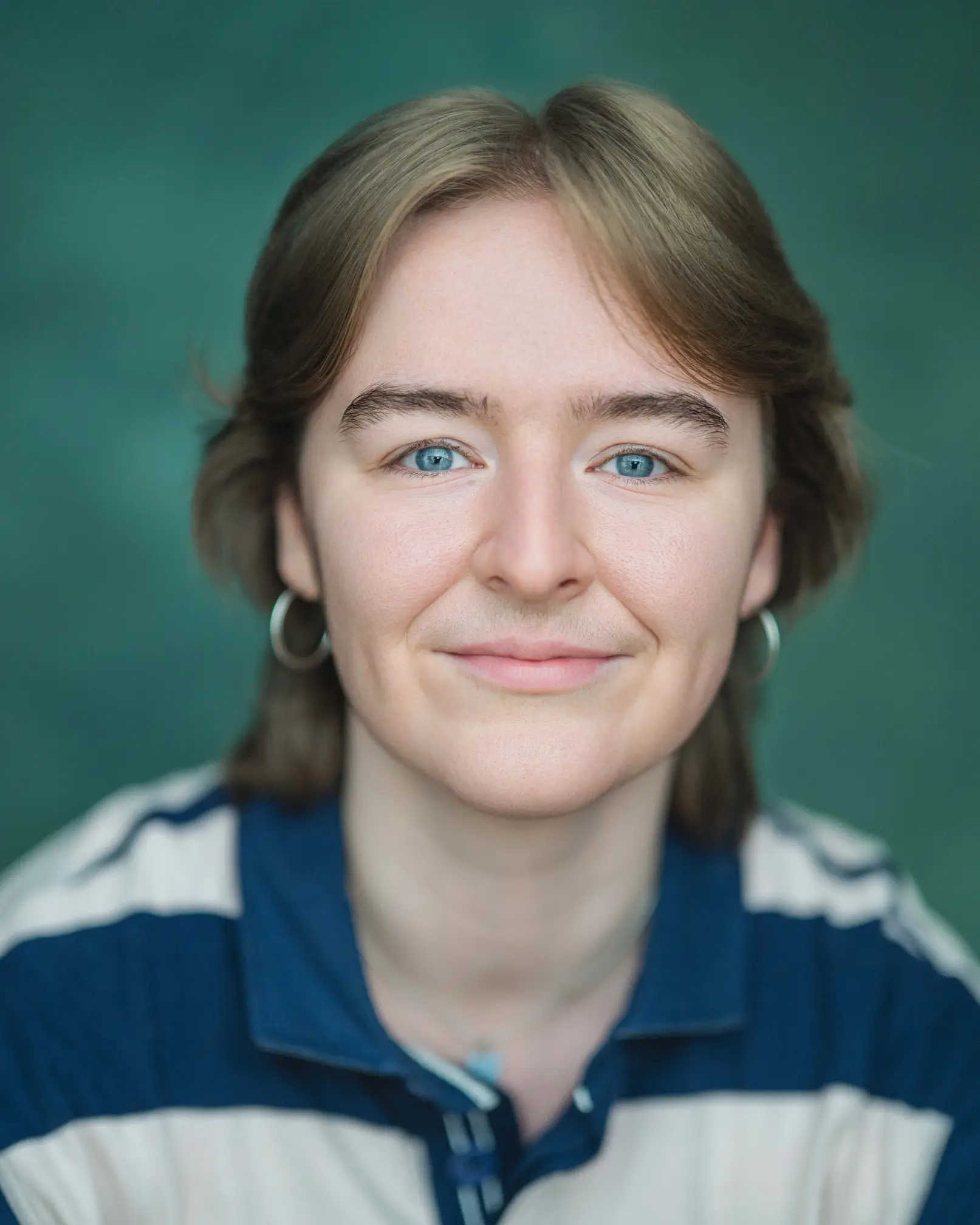 A white trans masc person with big blue eyes, shaggy short dark blond hair and stubble, wearing silver hoop earrings and a blue stripy rugby shirt against a dark green background.