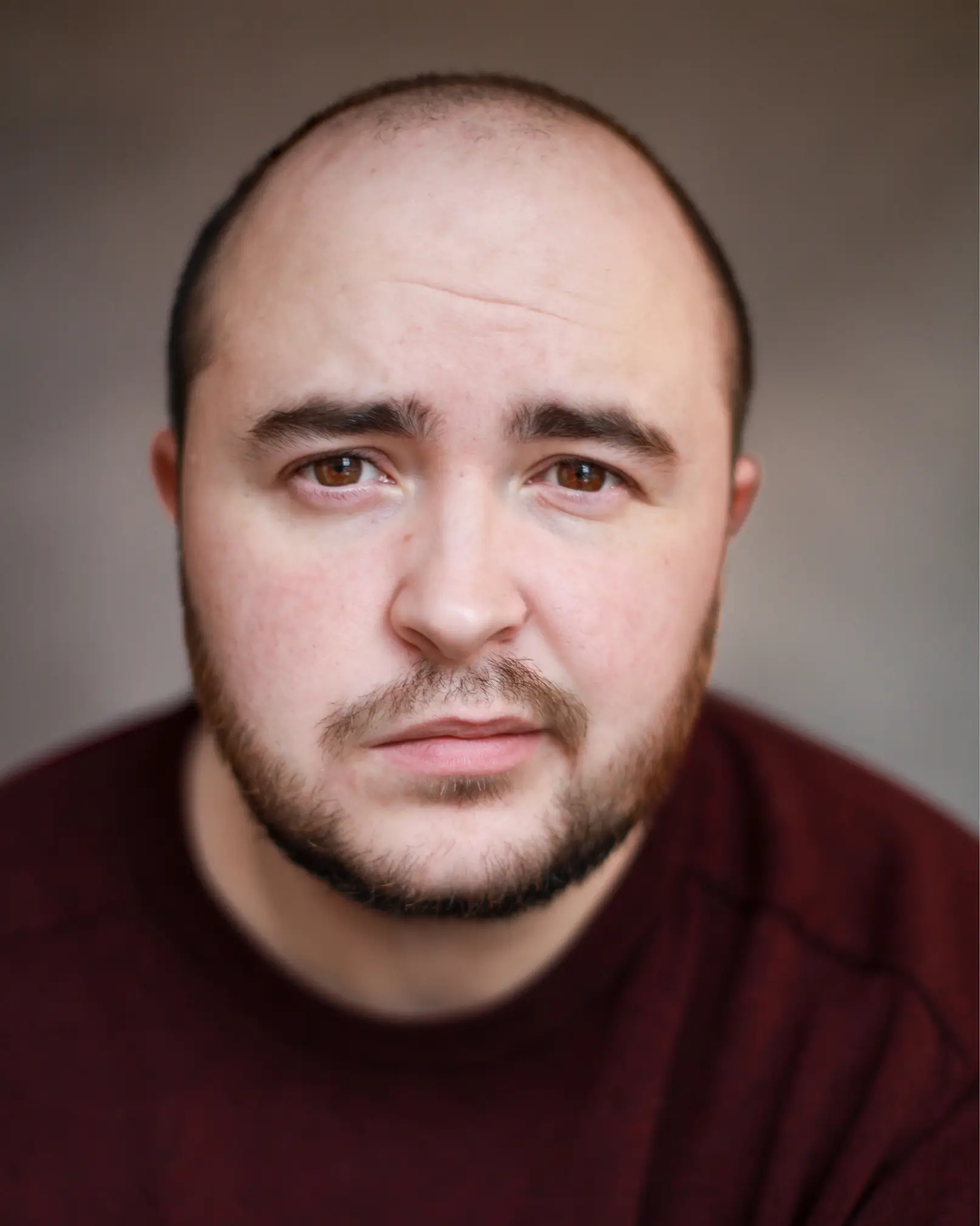 A headshot of a white trans man in his 20s with a shaved head and brown eyes. He has short brown and ginger stubble, and is wearing a burgundy jumper. He is looking directly at the camera.