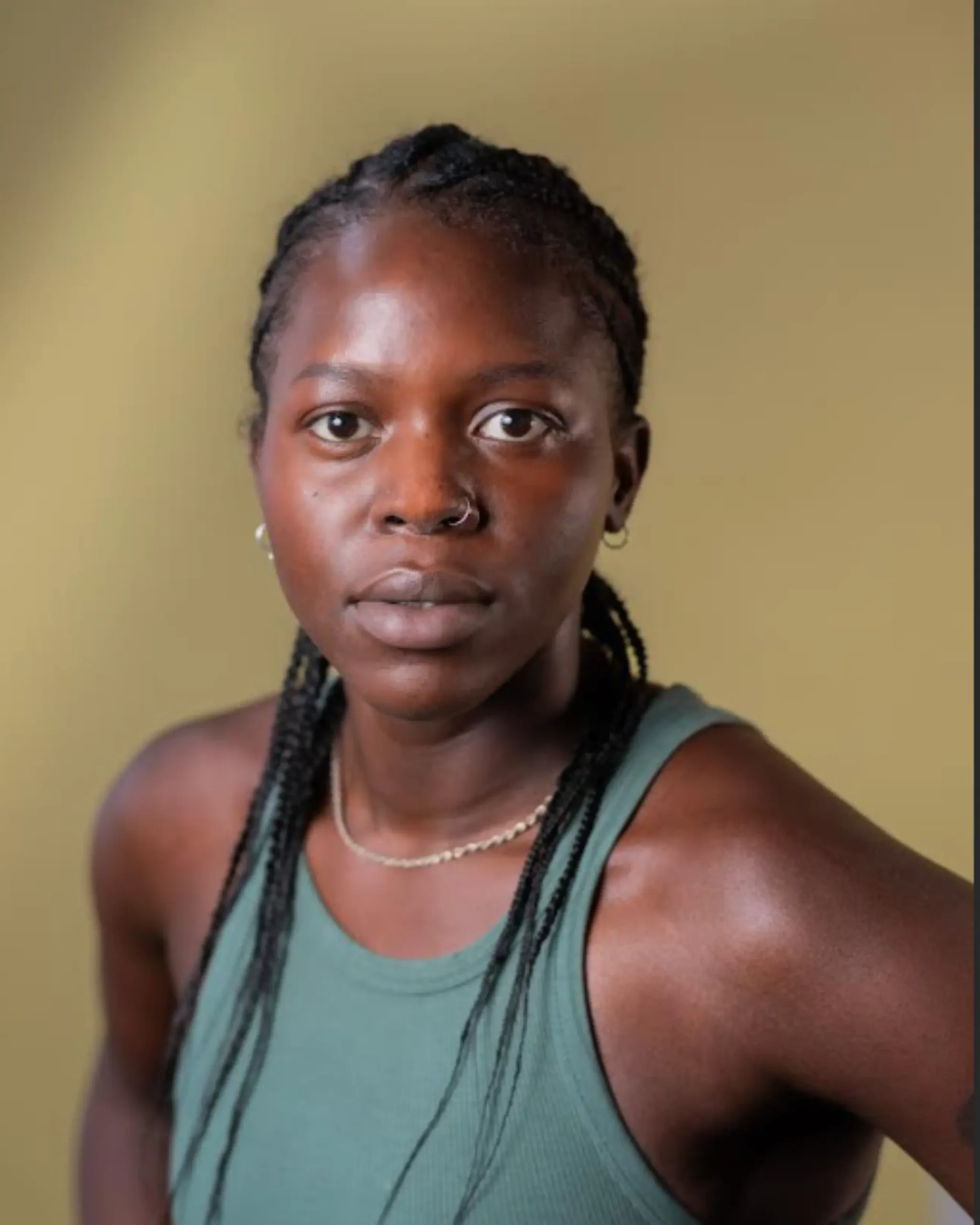 Headshot of Princess Bestman looking directly at the camera, in front of a blurred light green backdrop. Princess is are a Dark-Skinned Black person with nose peircings and braided hair. They're wearing a green ribbed vest and thin necklace.