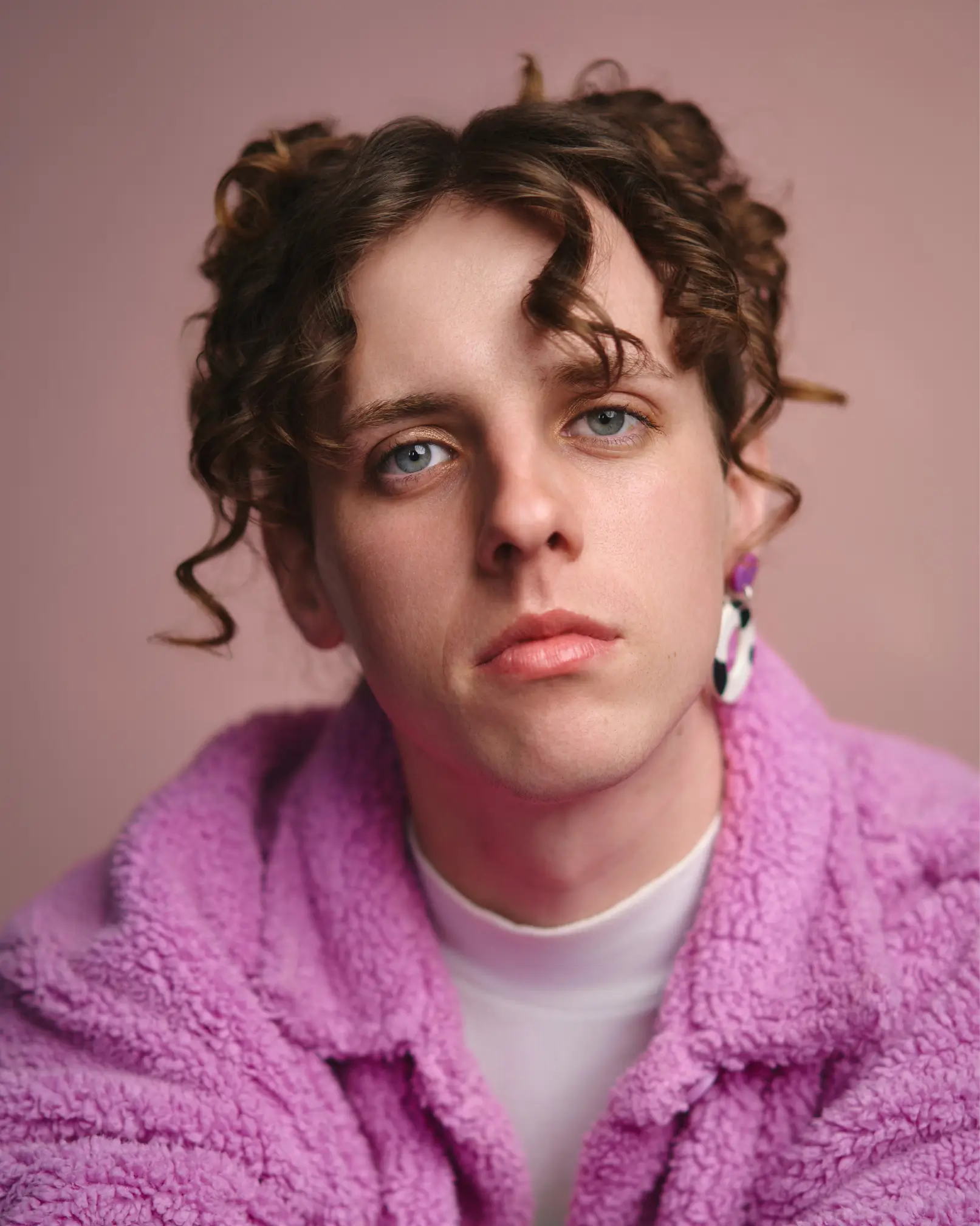 a white femme with brunette curly hair in messy spacebuns, wearing a fluffy pink coat and cow-print earring.