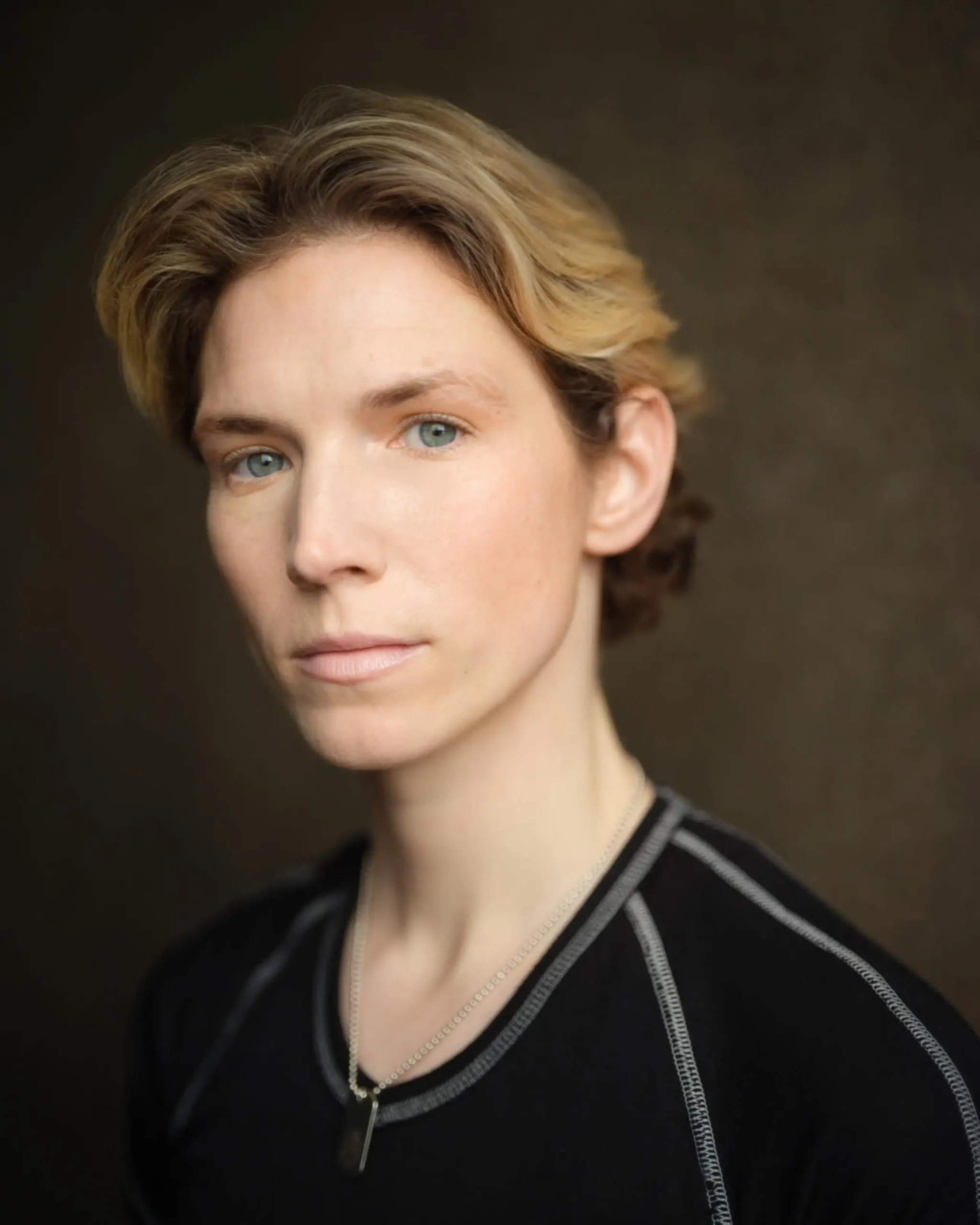 A white non binary person with short, blonde hair and light eyes looks directly to camera. They are wearing a black top and a silver necklace, set against a neutral background.