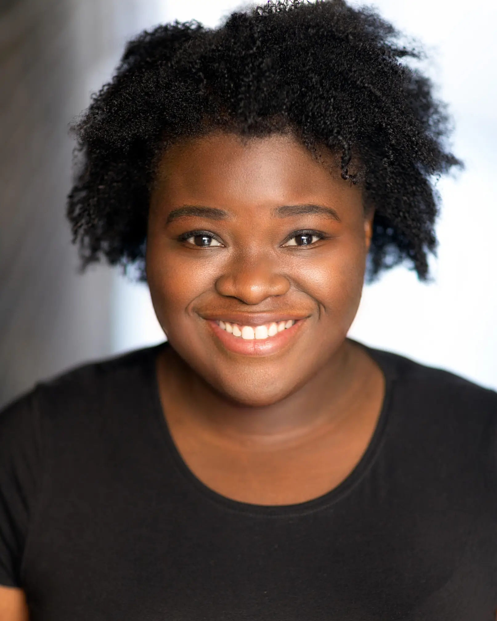 a dark-skinned Black femme with an oval face and dark eyes. They have long pink box braids and dark eyes. Their lips are a gradient of blue and purple, and they have a green to purple oil slick style of eyeshadow, encased by black graphic eyeliner. They are smiling and looking straight into the camera.